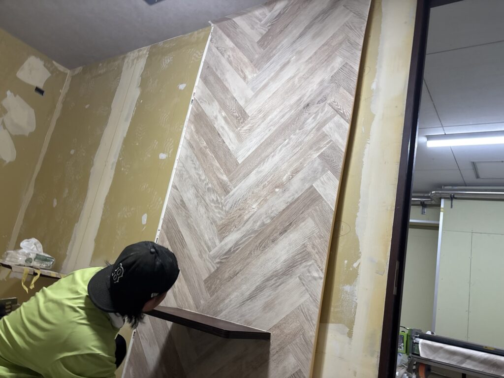The first strip of herringbone-patterned wallpaper being installed on a hotel room wall.