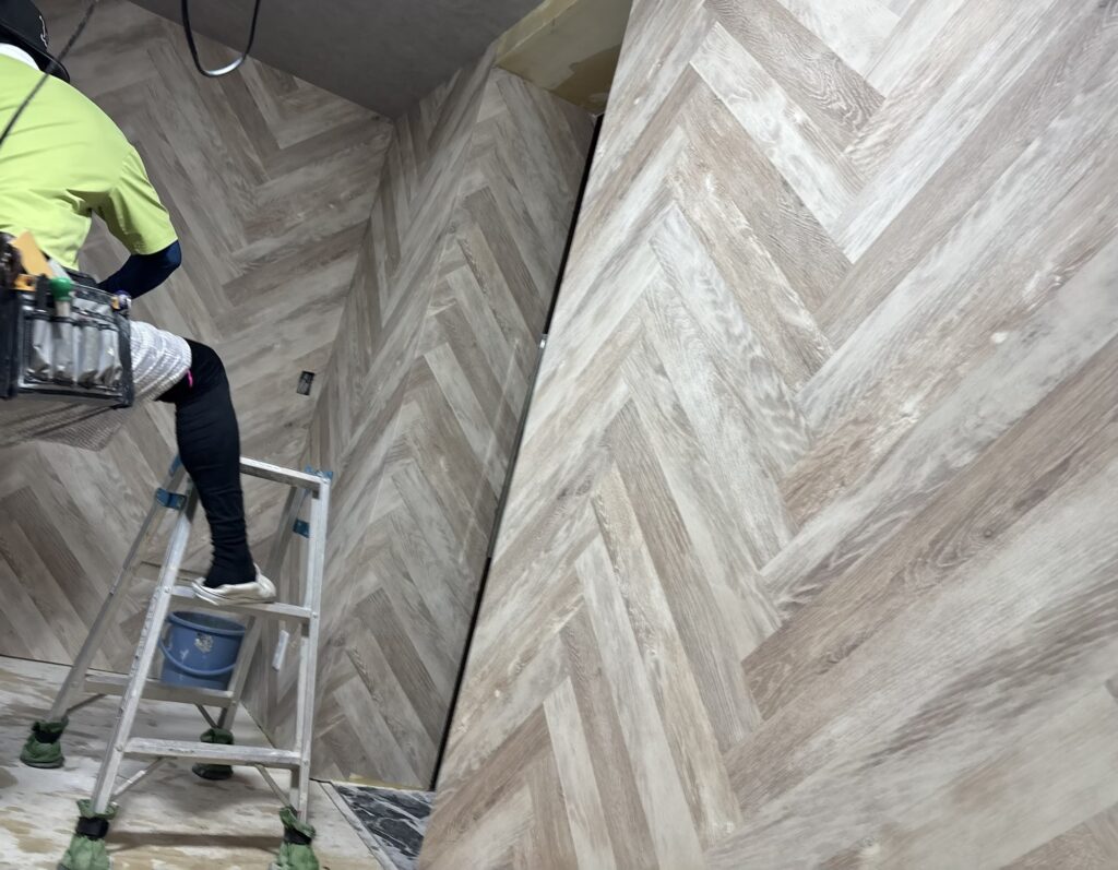 A hotel room wall with herringbone-patterned wallpaper almost fully installed.