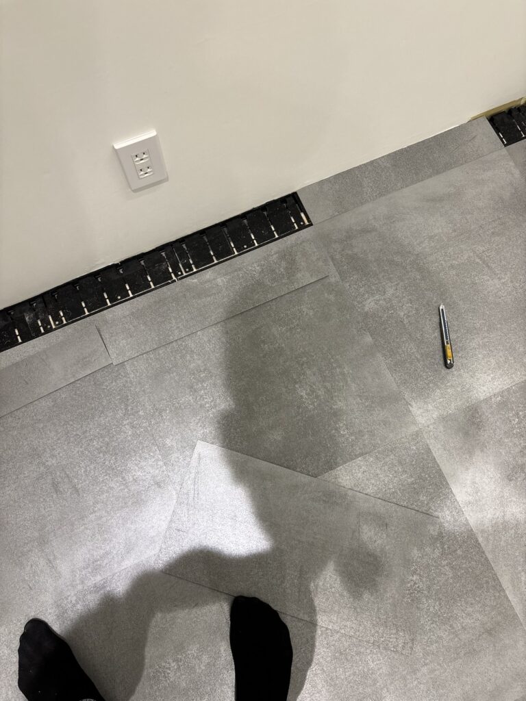 Worker installing gray vinyl floor tiles along the wall of a commercial store.