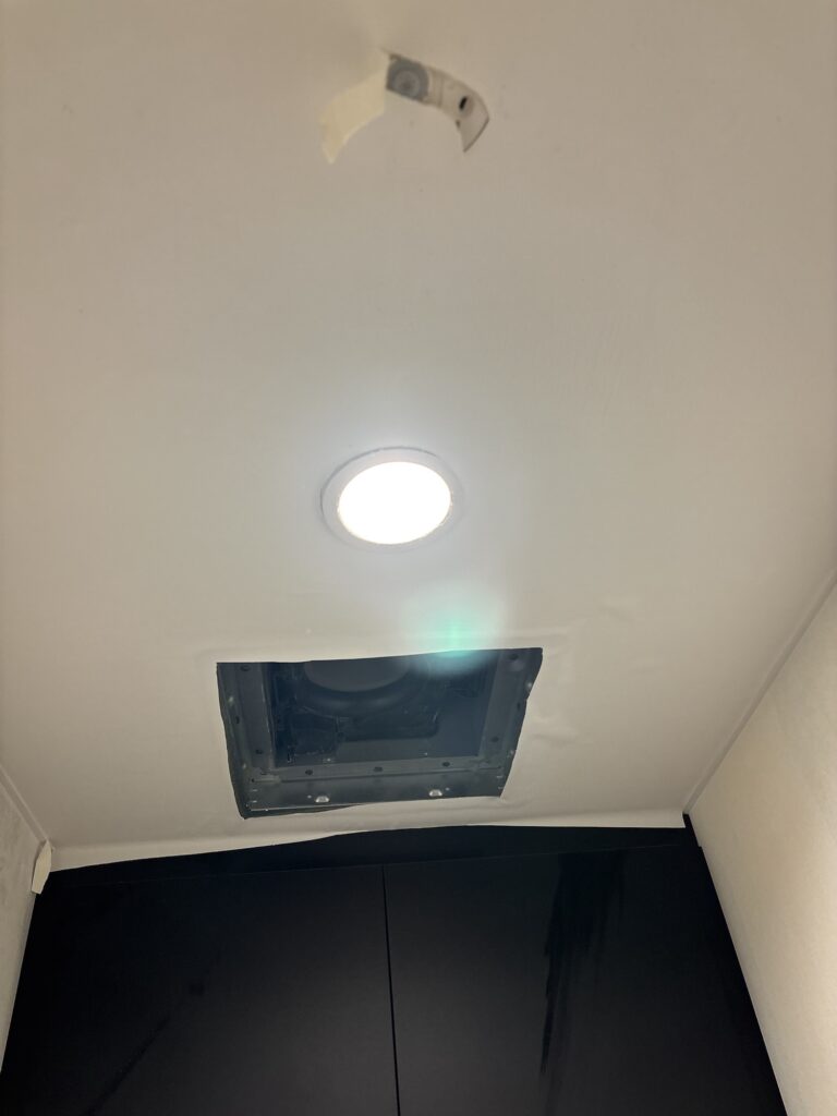 A worker installing white wallpaper on a toilet ceiling around a ventilation fan opening in a commercial renovation project in Japan.